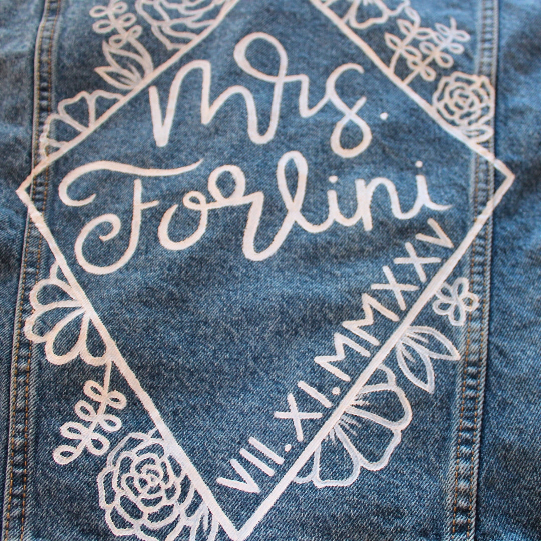 Hand-Painted Bridal Denim Jacket featuring delicate flowers, personalized with initials and love quote. Unique and chic fashion for a memorable wedding day. #BridalStyle #WeddingFashion