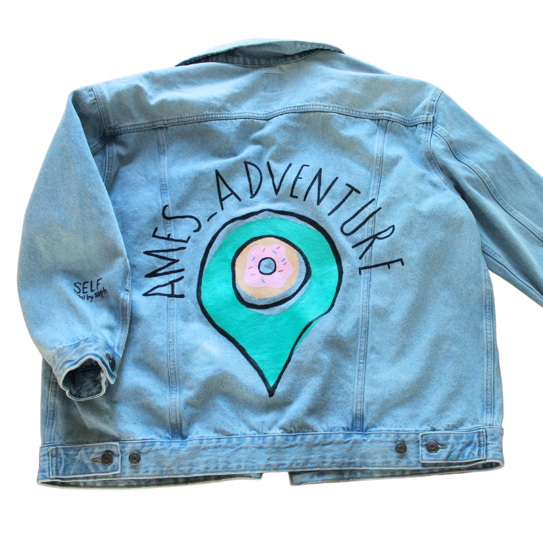 A denim jacket with a prominently displayed logo hand painted on the back of the jacket.