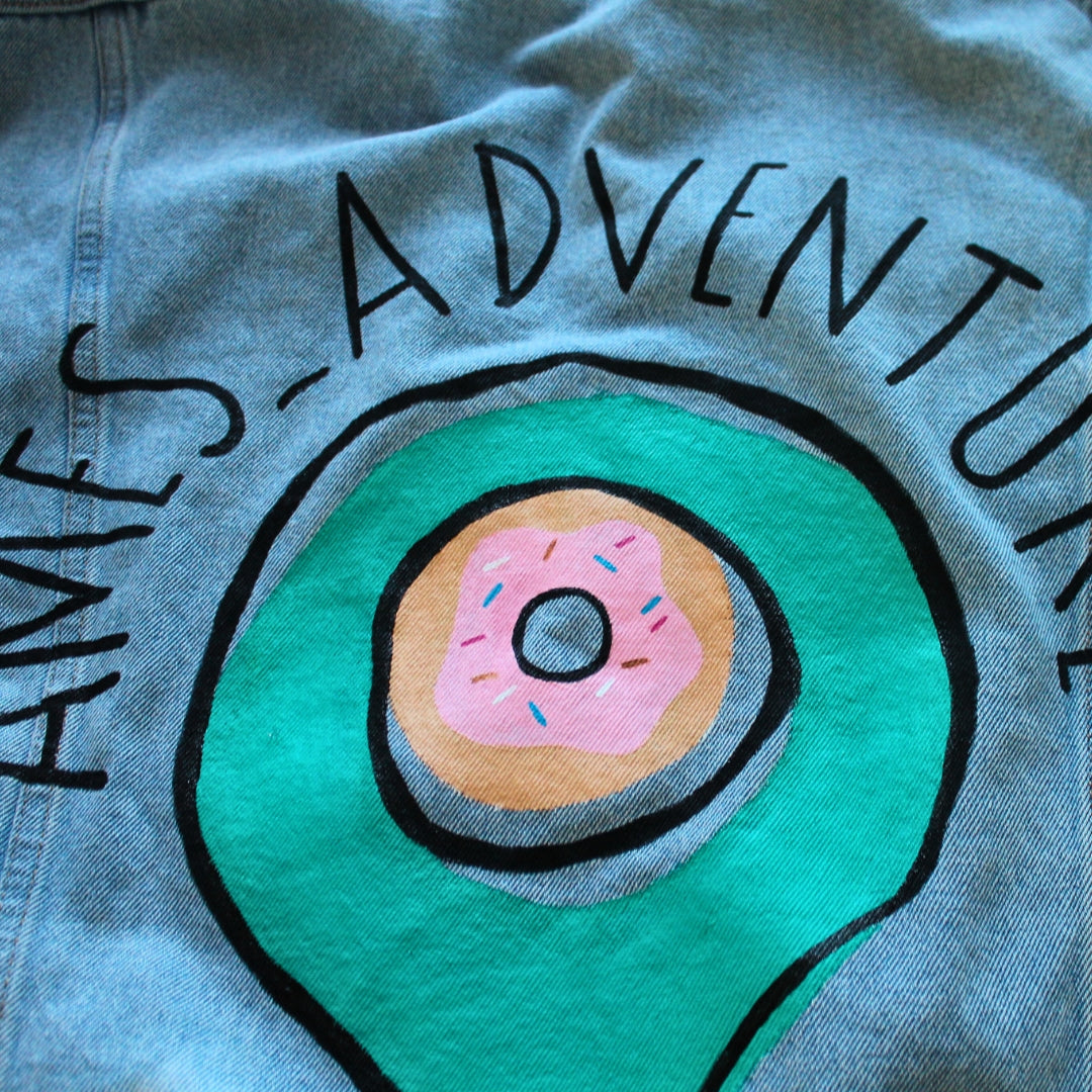 A denim jacket with a prominently displayed logo hand painted on the back of the jacket.